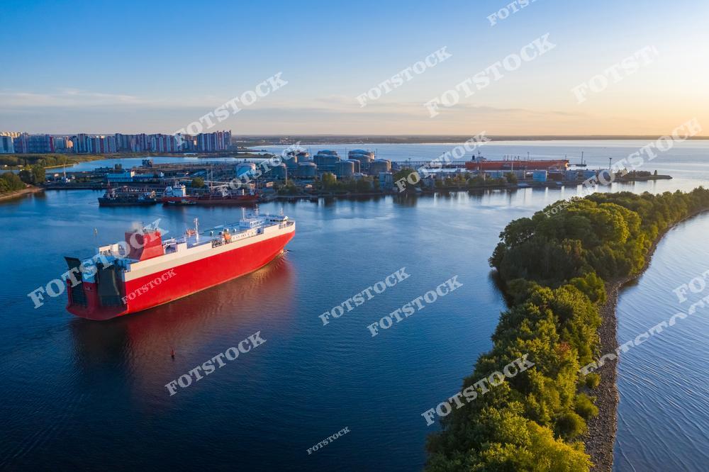 Фотография показывает грузовой корабль, находящийся в акватории морского порта. На заднем плане видны промышленные здания и жилой район, а зеленая растительность набережной создает контраст. Снимок демонстрирует взаимодействие природы и человеческой деятельности.